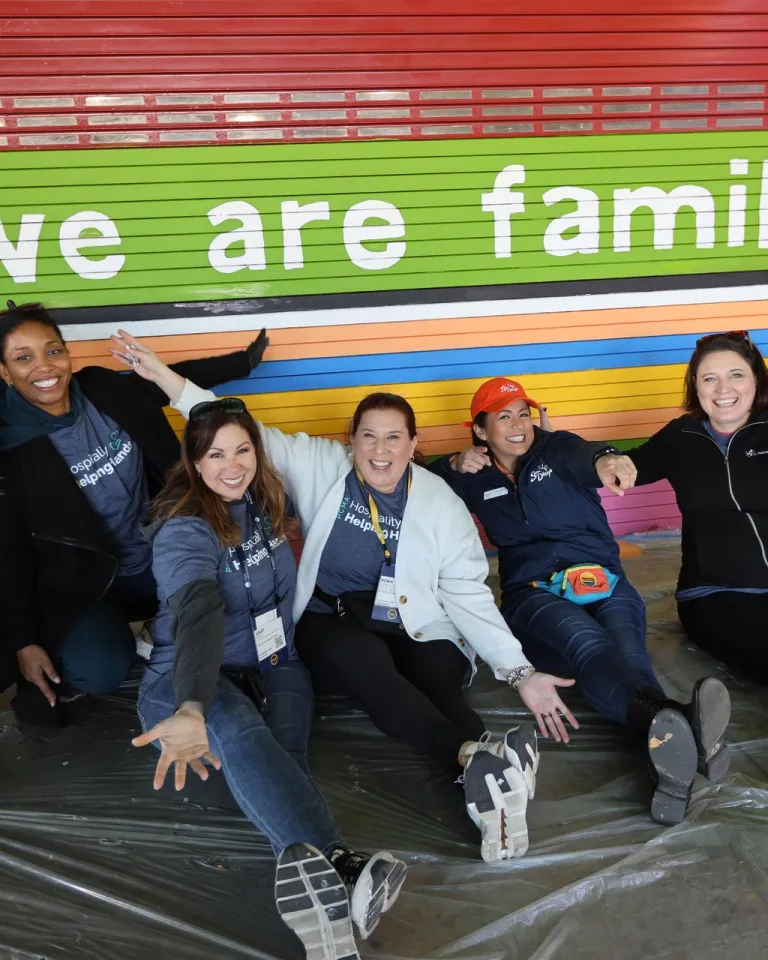 A group of seven smiling adults in matching event T‑shirts sit on a plastic-covered floor in front of a brightly colored mural in San Diego that reads ‘we are family’ with a small globe icon. They pose playfully with arms raised and legs stretched toward the camera, appearing energetic and celebratory during a community engagement event benefiting a local grade school.