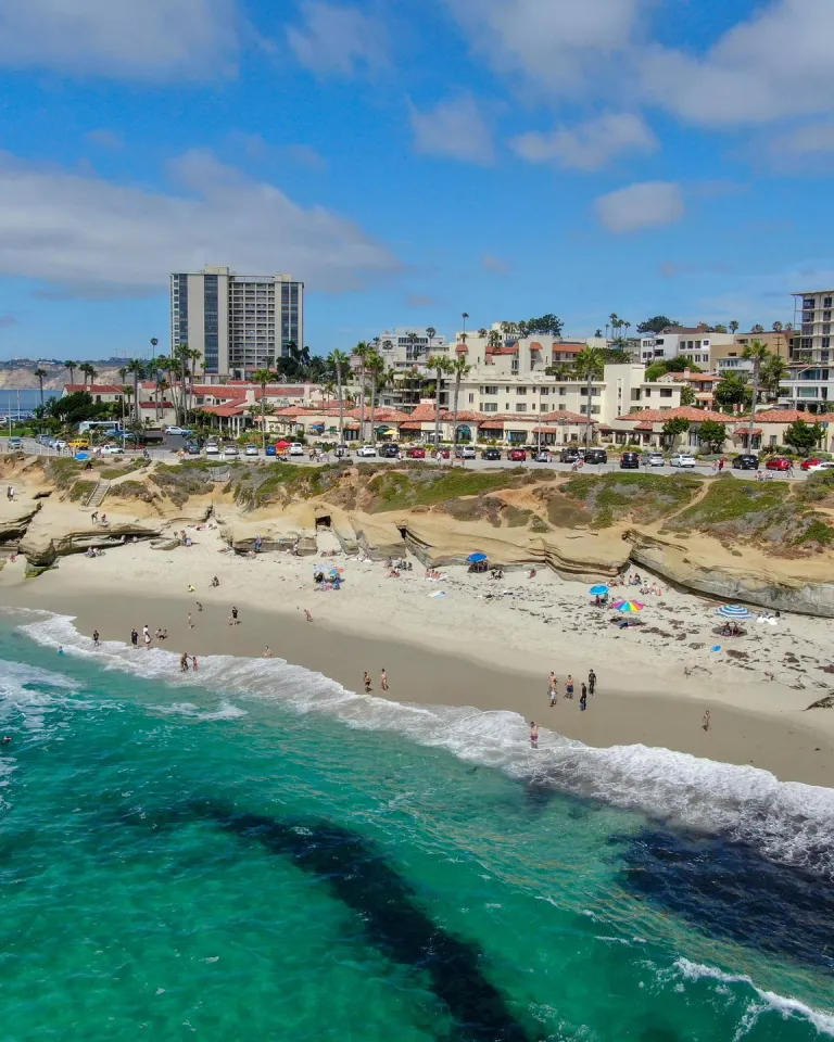 Aerial view of La Jolla Cove in San Diego, showing turquoise ocean waters, a sandy beach with visitors, rugged coastal cliffs, and nearby buildings under a bright blue sky.