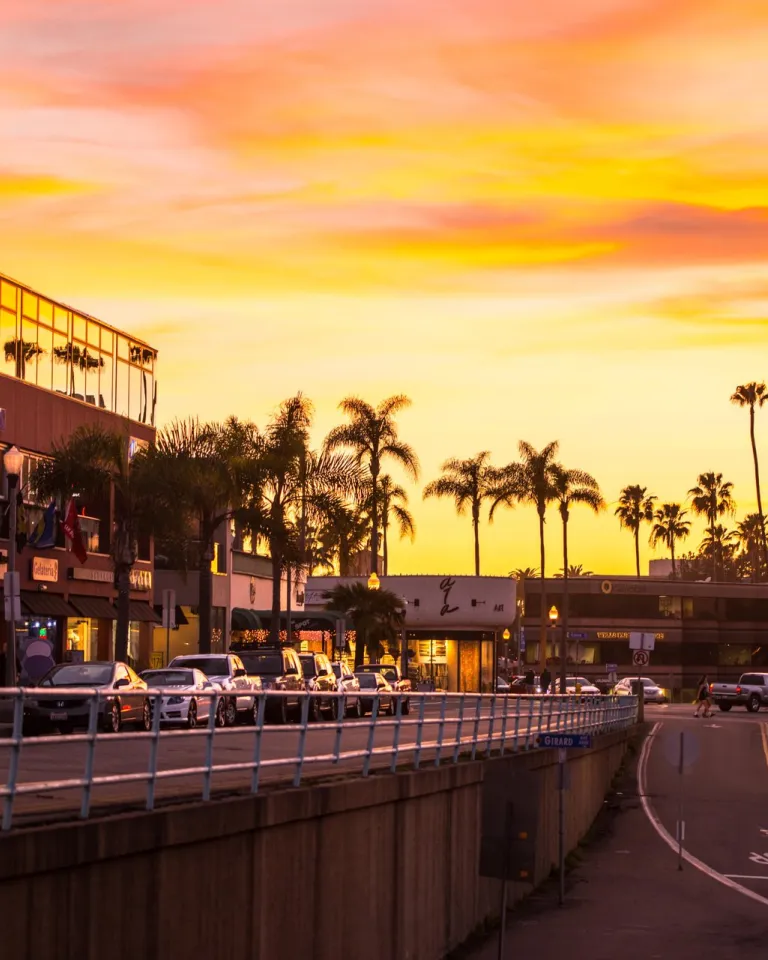 Sunset view of the Village of La Jolla in San Diego, with palm-lined streets, coastal shops and restaurants, light traffic, and a warm orange and pink sky reflecting a vibrant seaside atmosphere.