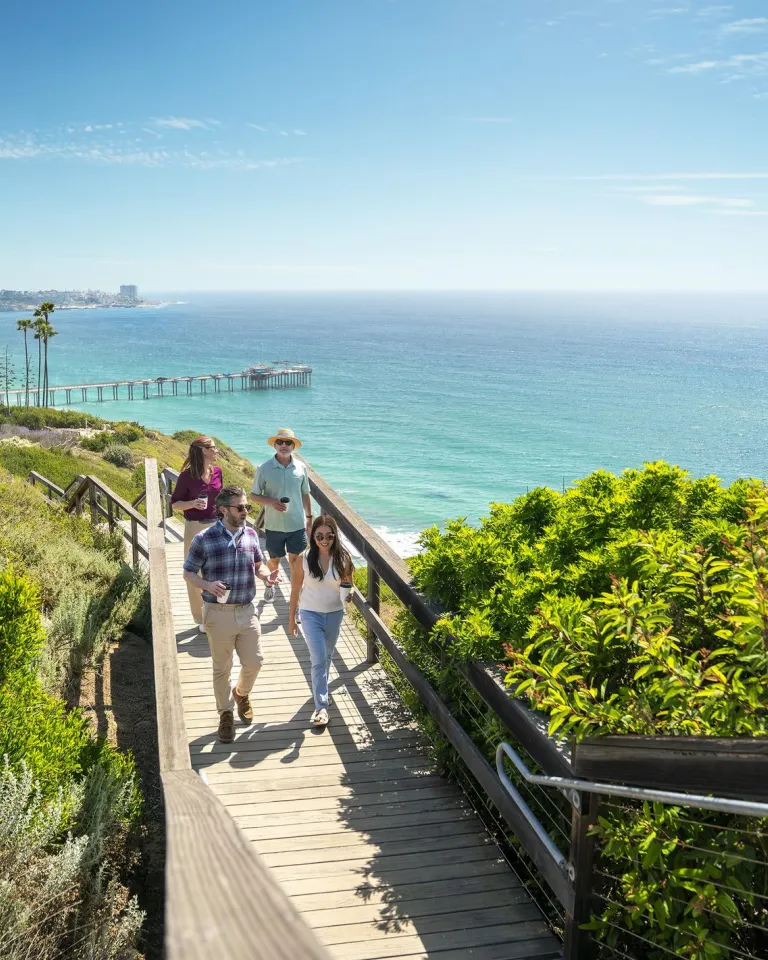 Four people in business casual attire stroll on a wooden walkway at the Scripps Coastal Meander in the La Jolla community of San Diego, against the backdrop of a sparkling blue-green sea and sunny blue skies.