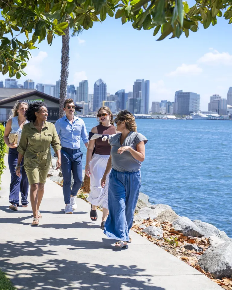 Five people walk along a waterfront sidewalk under sunny skies on Shelter Island, with the beautiful San Diego skyline in the background.