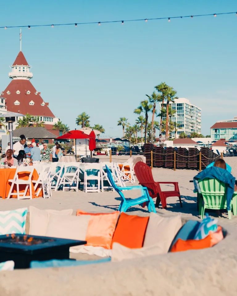 A view of the Hotel del Coronado in the beautiful San Diego region, depicting people lounging in beach chairs on the sand against a backdrop of blue skies and the hotel's brick-red spires.