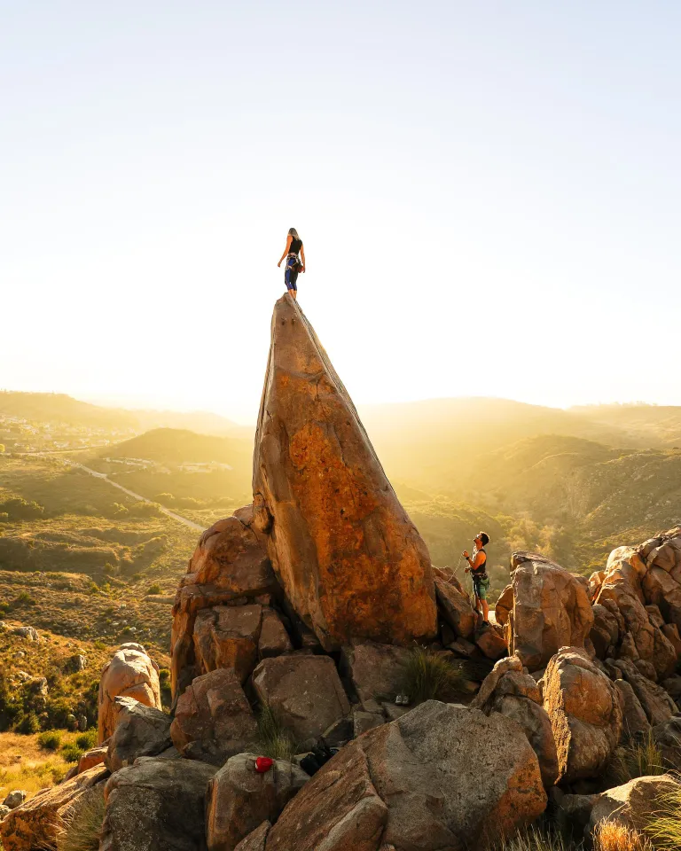 Hikers standing on a dramatic rock formation in San Diego, overlooking rolling hills and open landscapes during golden hour with warm sunlight illuminating the terrain.