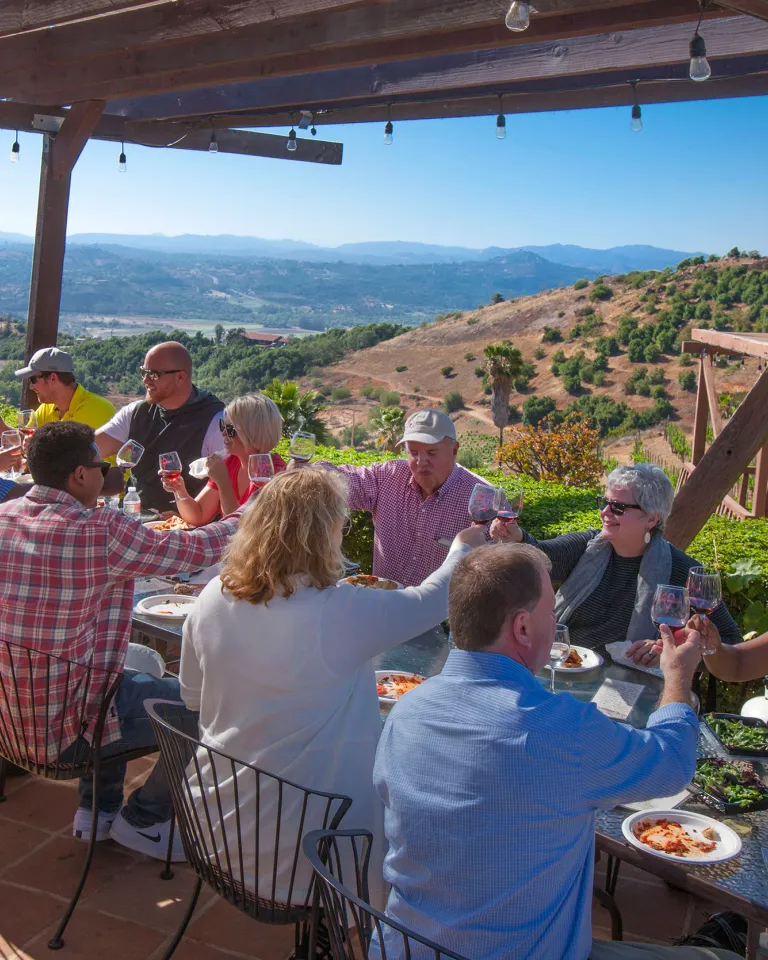 Group meeting taking place on an outdoor patio in San Diego's North Inland area, with attendees seated around a table enjoying food and conversation while overlooking rolling hills and a scenic valley.