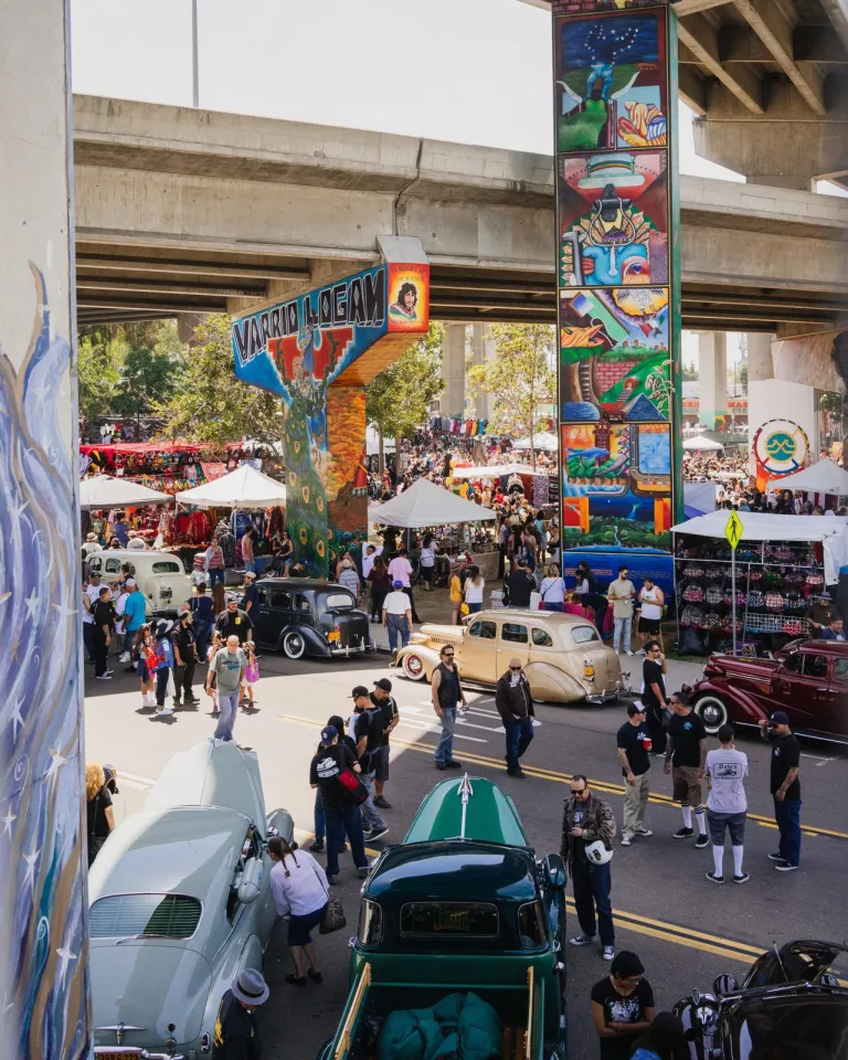 A vibrant street festival at Chicano Park in Barrio Logan under an overpass, featuring classic cars, food stalls, and colorful murals.