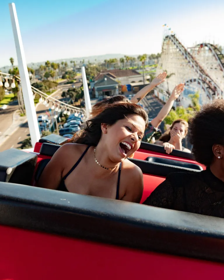 Two young women enjoy the Giant Dipper roller coaster ride at Belmont Park, laughing against a bright, sunny backdrop.