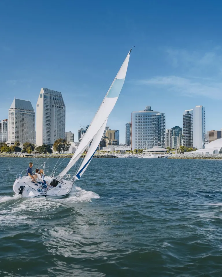 A sailboat glides across San Diego Bay under blue skies on a beautiful day, with the Downtown San Diego skyline in the background.