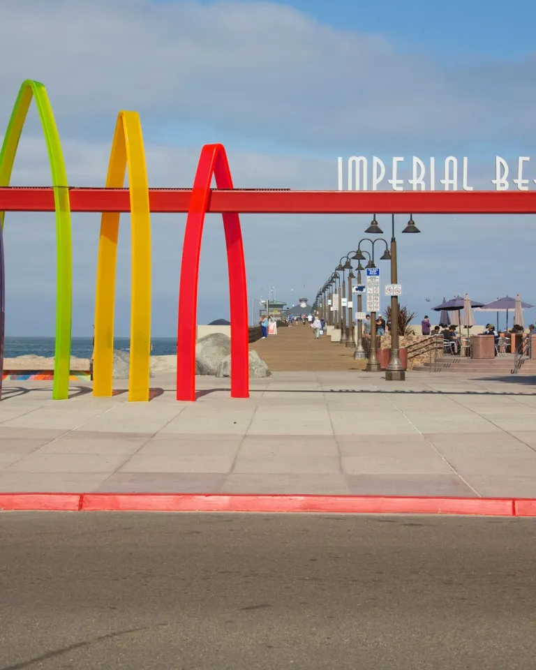A colorful gateway and sign reading "Imperial Beach" stands at a beachfront, with the entrance to the Imperial Beach Pier near San Diego in the background.