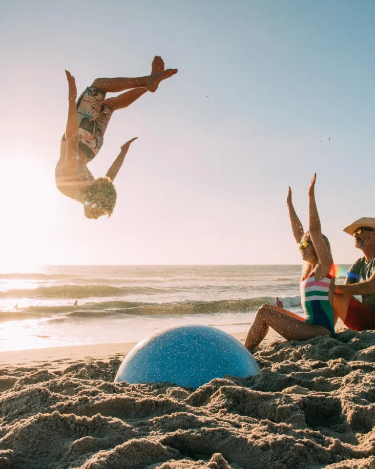 A child flips in the air above a San Diego beach, while two adults cheer on the sand at sunset.