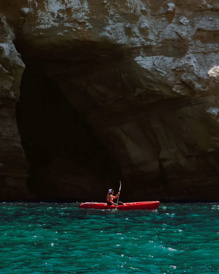 Alana Nichols in a red kayak paddles near a rocky cliff and a cave entrance in turquoise water.