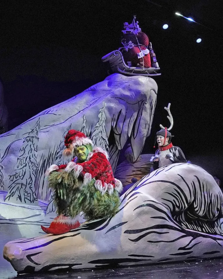 The Grinch in a Santa outfit sits on a snowy set, with characters and colorful lights.