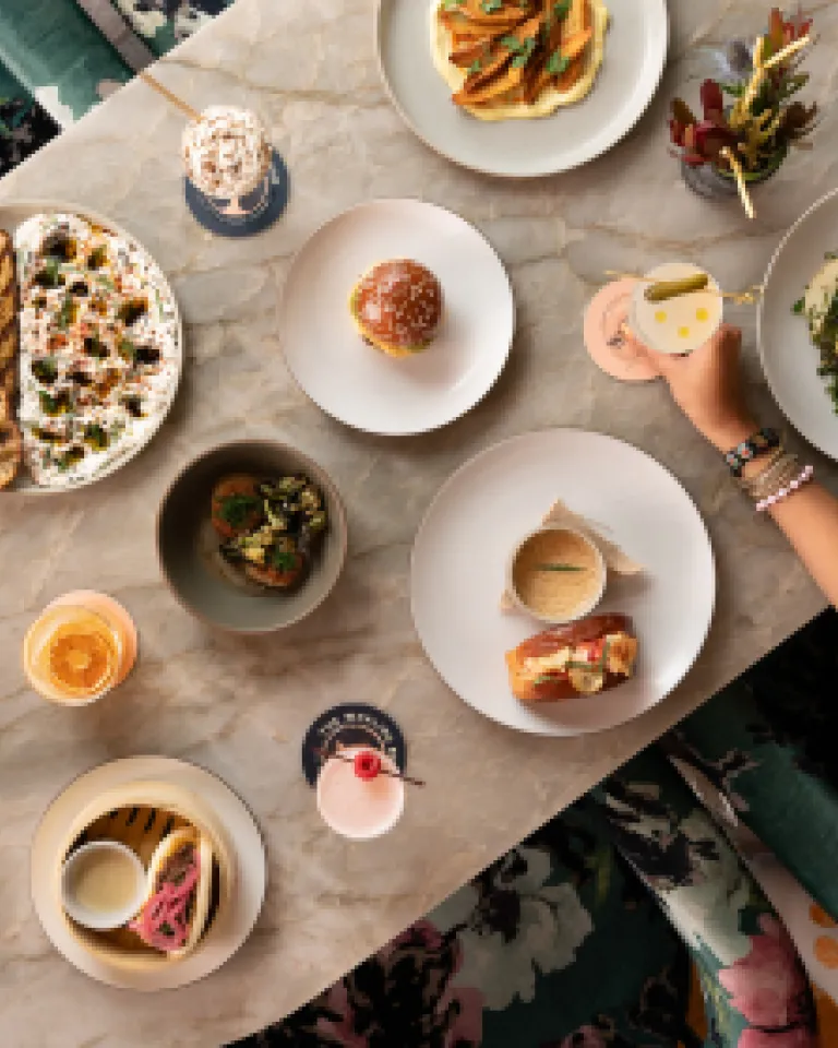 A tabletop spread featuring various dishes, drinks, and a hand reaching for food.