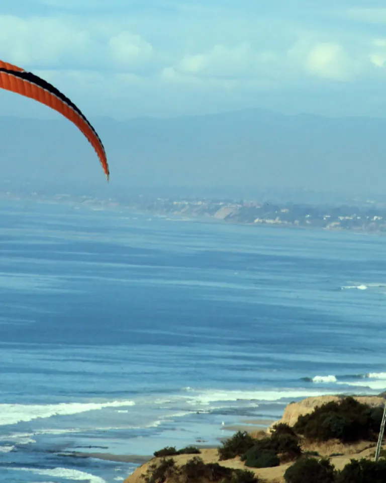 A paraglider soars over the coastal bluffs of San Diego, with the Pacific ocean in the background.