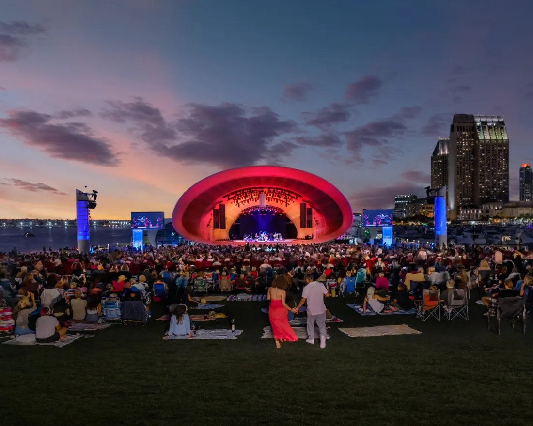 A large crowd gathers on a lawn facing an outdoor concert stage illuminated in red, with city buildings and a sunset sky in the background.