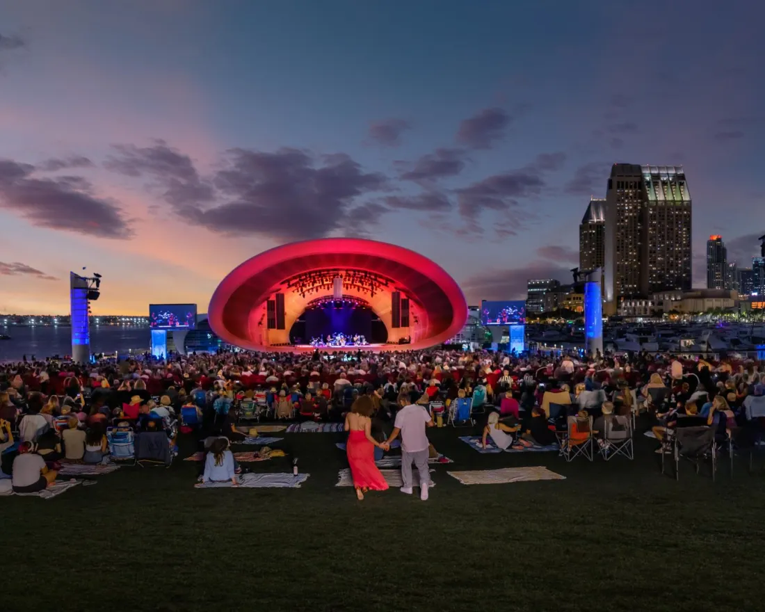 A large crowd gathers on a lawn facing an outdoor concert stage illuminated in red, with city buildings and a sunset sky in the background.