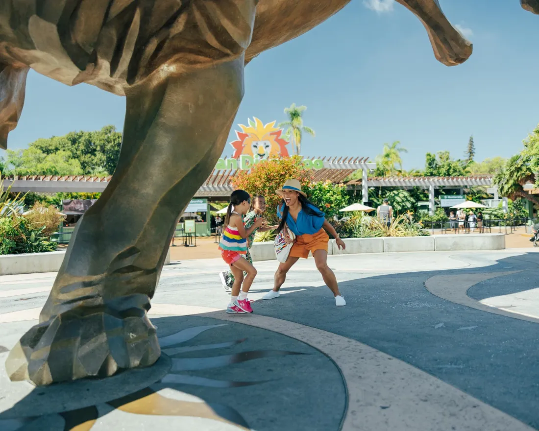 Two people pose playfully under a large dinosaur statue at a zoo or amusement park on a sunny day.