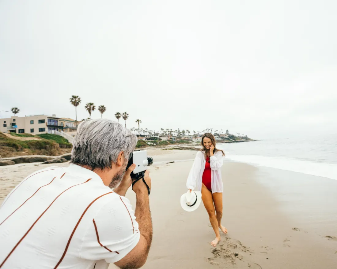 A man takes a photo of a woman posing on a sandy San Diego beach near the ocean. The woman holds a hat and smiles, and palm trees and houses are visible in the background.