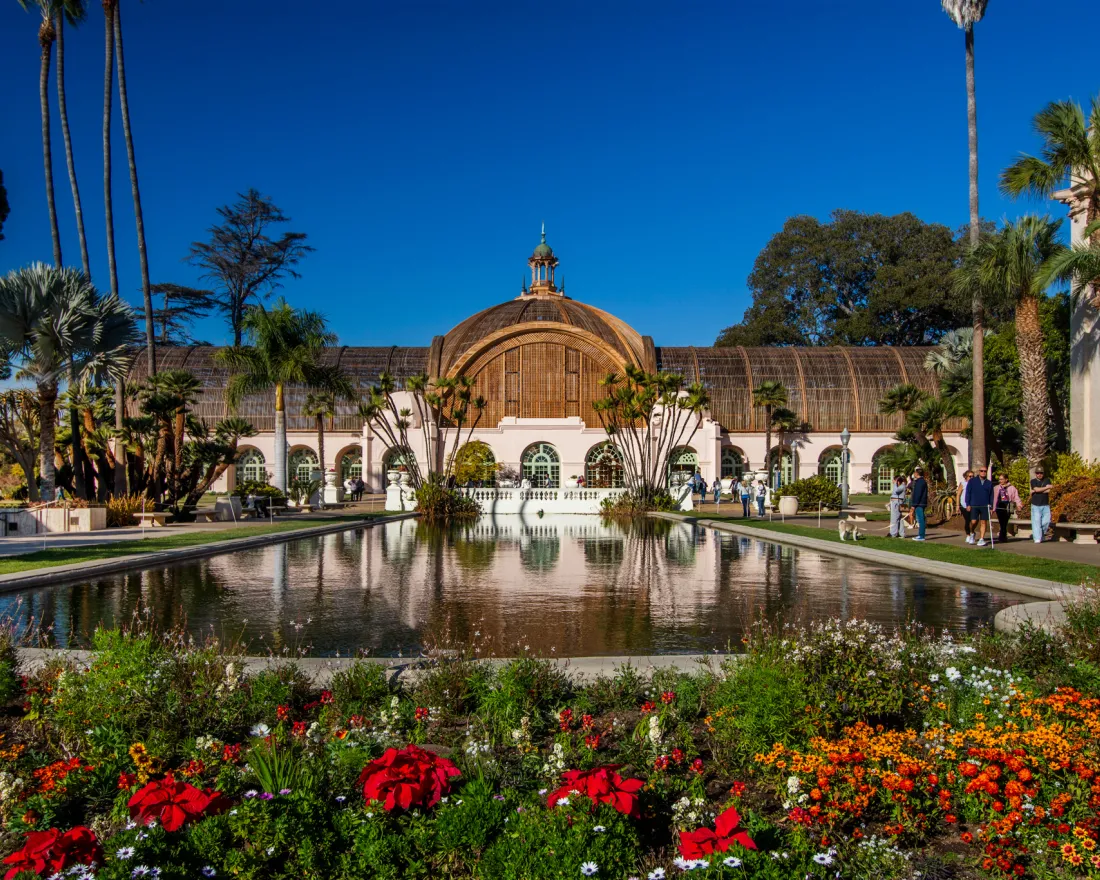 Balboa Park Botanical building with arched windows behind a reflective pond, surrounded by colorful flower beds, palm trees, and visitors on a clear day.