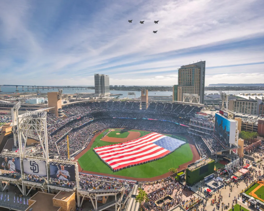 A large American flag is displayed on petco park, with four jets flying in formation overhead and a crowd filling the stands.
