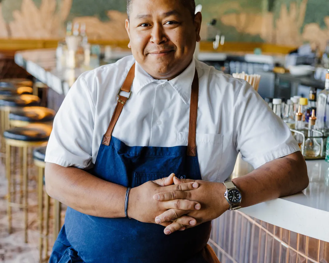 A chef wearing a white shirt and blue apron, David Sim, stands behind a bar counter in a restaurant, smiling with hands clasped.