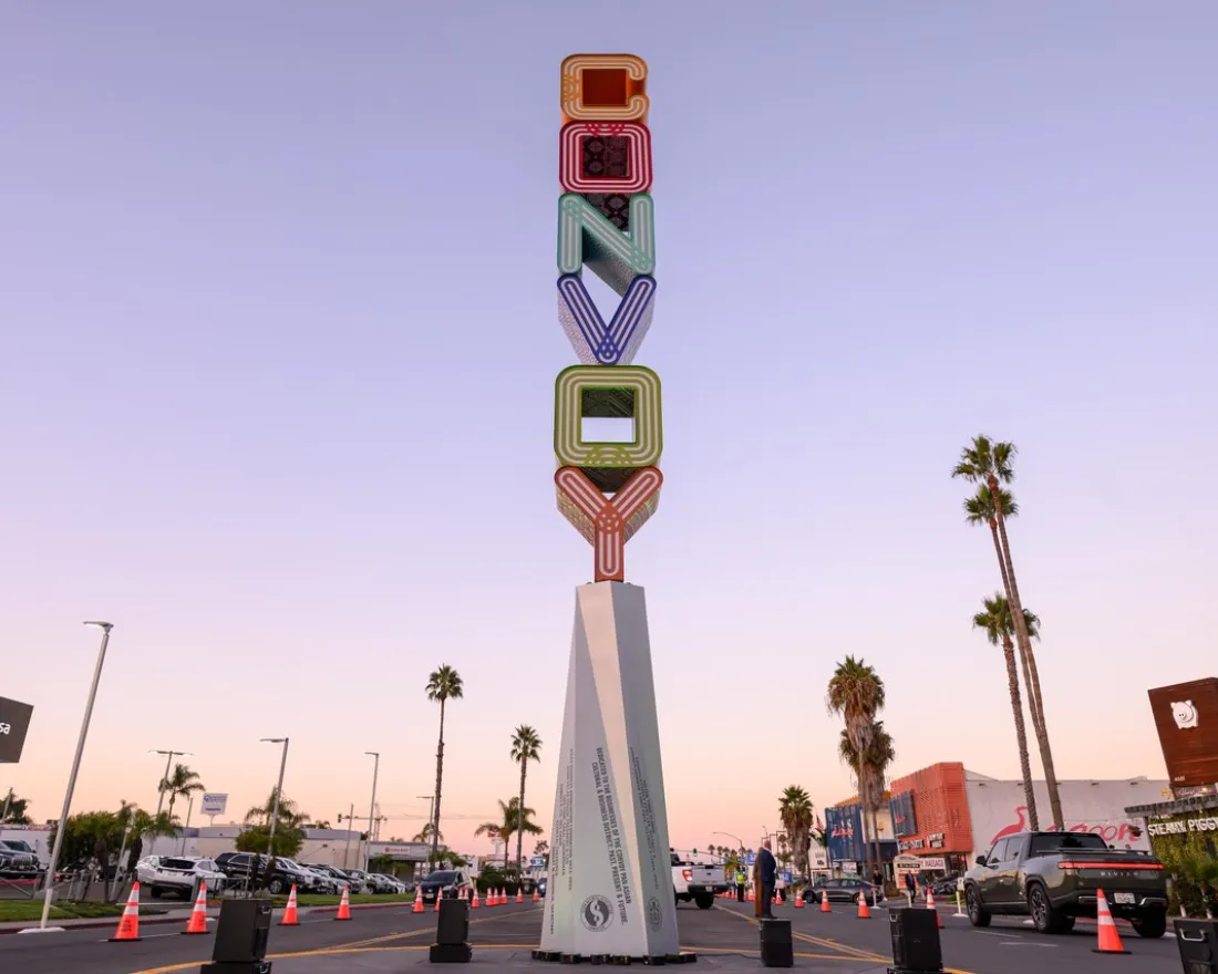 A tall vertical sign spelling "Convoy" in colorful letters stands in the middle of a street lined with palm trees, cars, and shops at sunset.