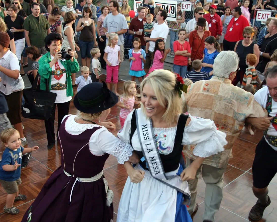 People in traditional clothing dance at an outdoor Oktoberfest in San Diego while a crowd of onlookers, including children, watch. A woman wearing a "Miss La Mesa" sash is smiling in the center.
