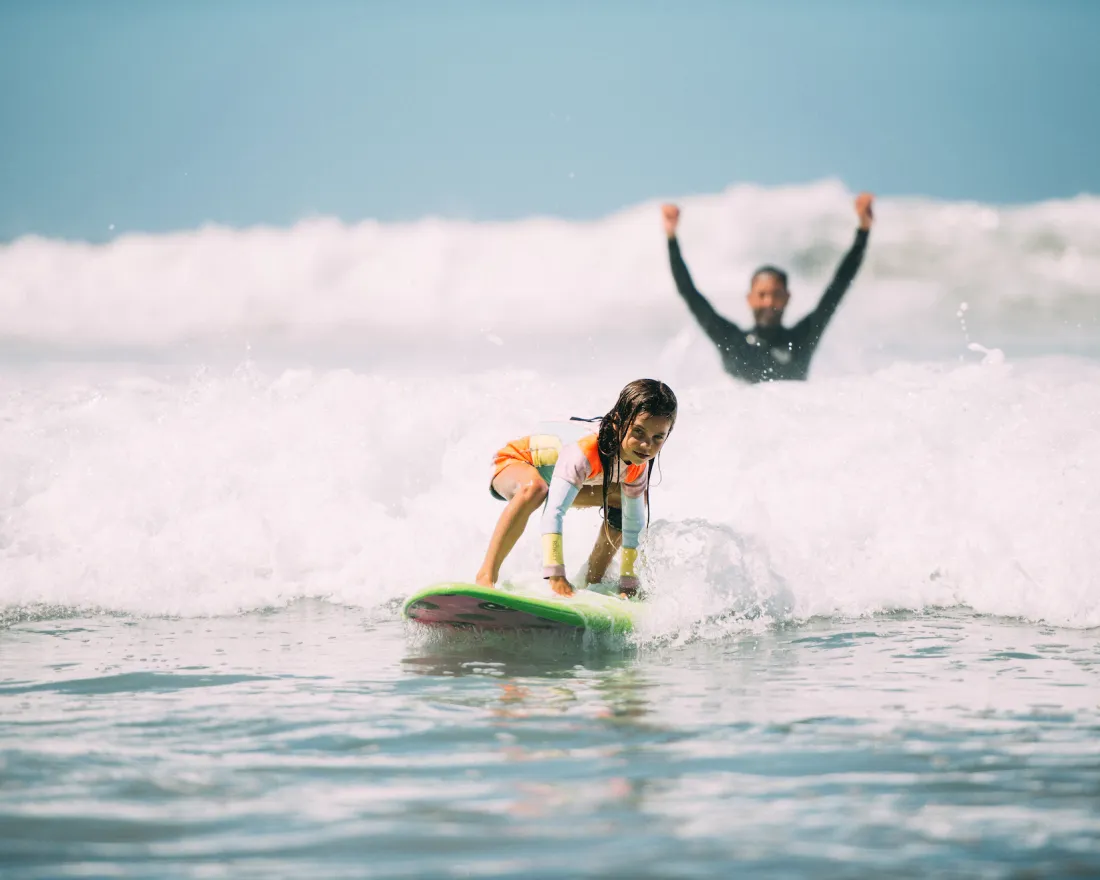 A young girl begins to stand up on a surfboard in the whitewater of a wave on a sunny day at a San Diego Beach, as her instructor raises his arms joyously behind her.