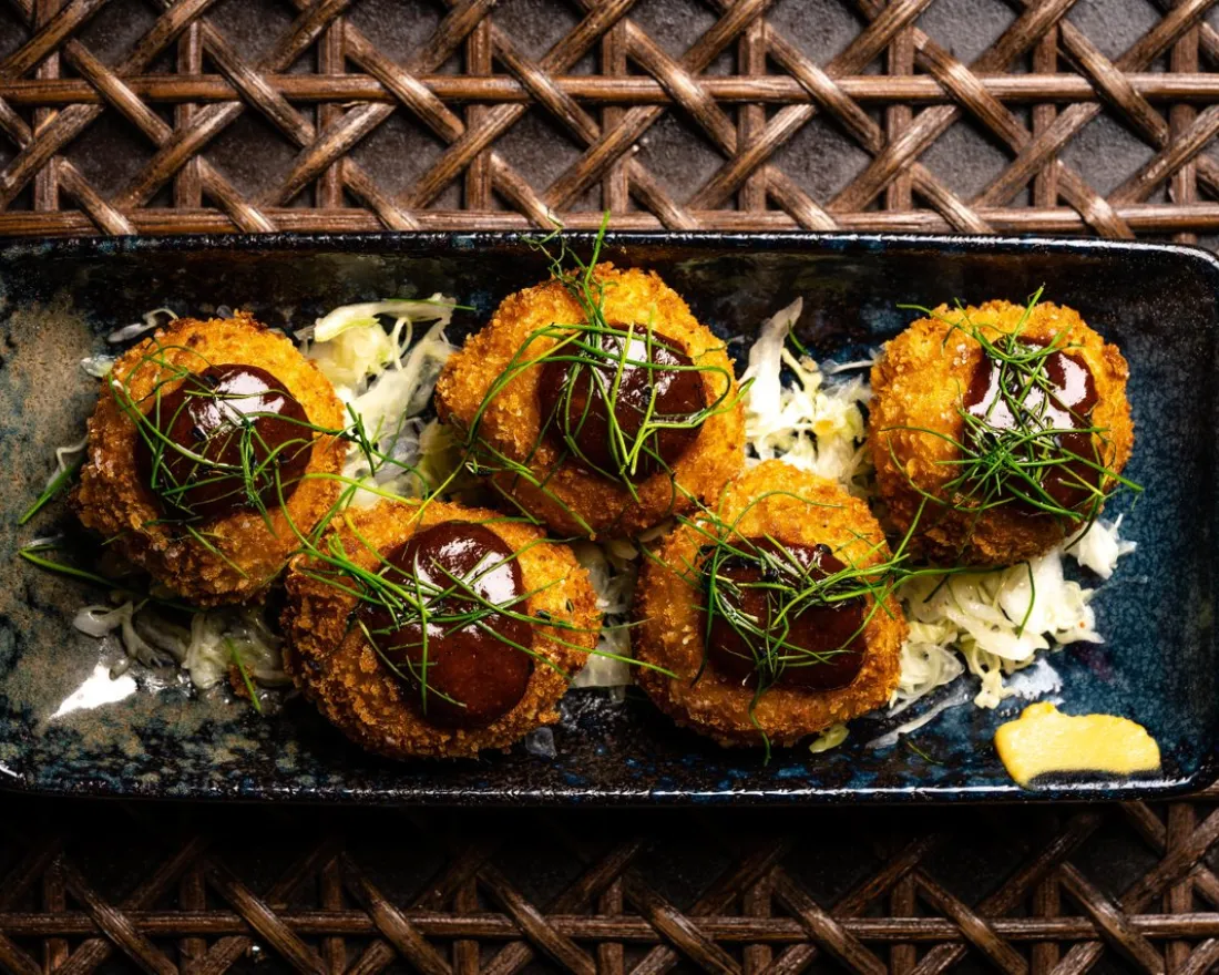 Six breaded, fried croquettes topped with sauce and herbs are arranged on shredded cabbage in a rectangular blue plate at Realm of the 52 Remedies in San Diego.