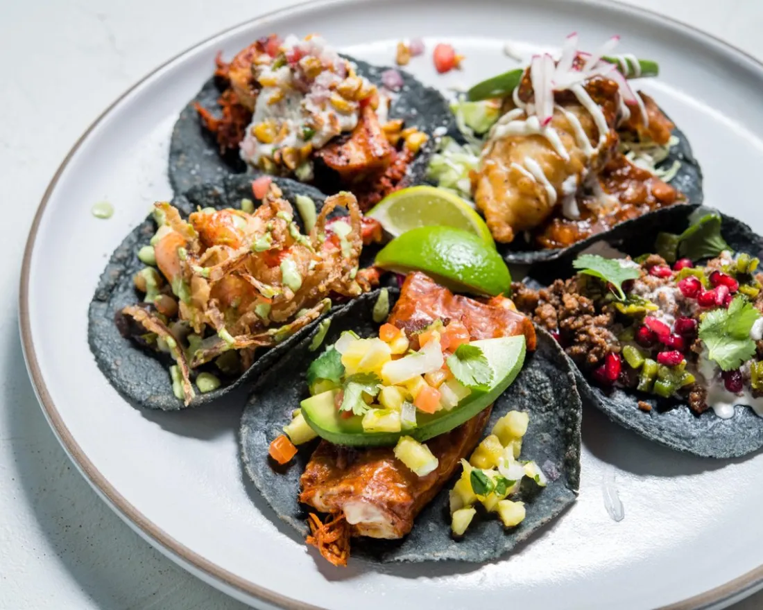A plate with five assorted tacos from Puesto on blue corn tortillas, topped with various meats, vegetables, sauces, and garnishes, with lime wedges in the center.