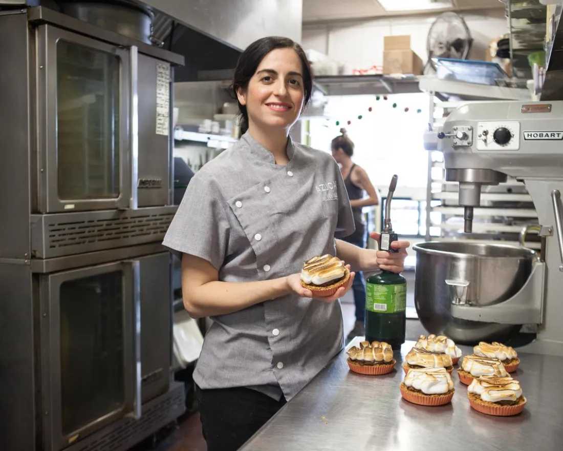 Chef Vivian Hernandez of Azucar, in a gray uniform, stands in a commercial kitchen, holding a tart and a whip cream canister, with several finished tarts on the counter beside her.