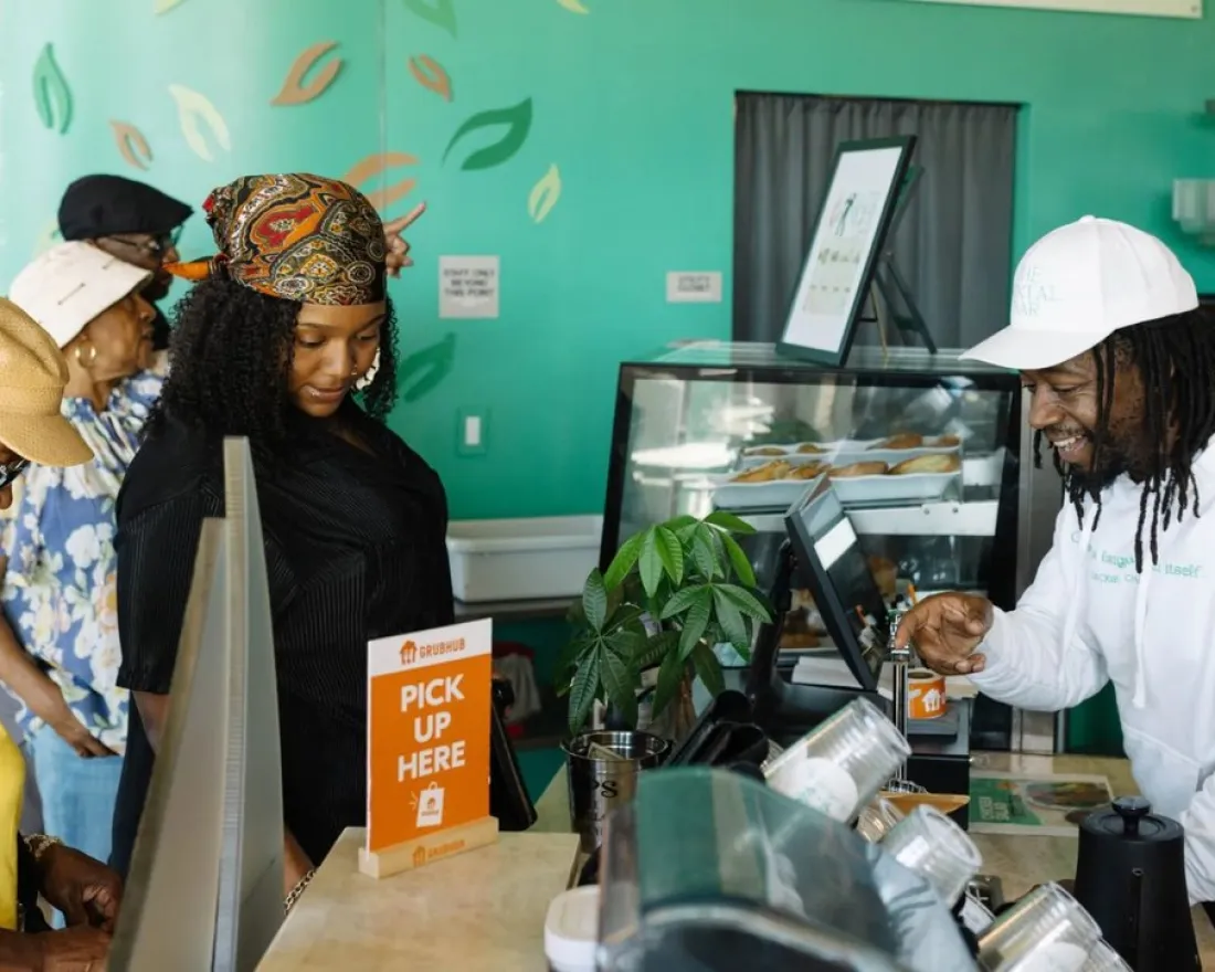 Customers wait at the counter at The Mental Bar in San Diego while a staff member in a white hoodie and cap assists them in a brightly decorated cafe with a "Pick Up Here" sign.