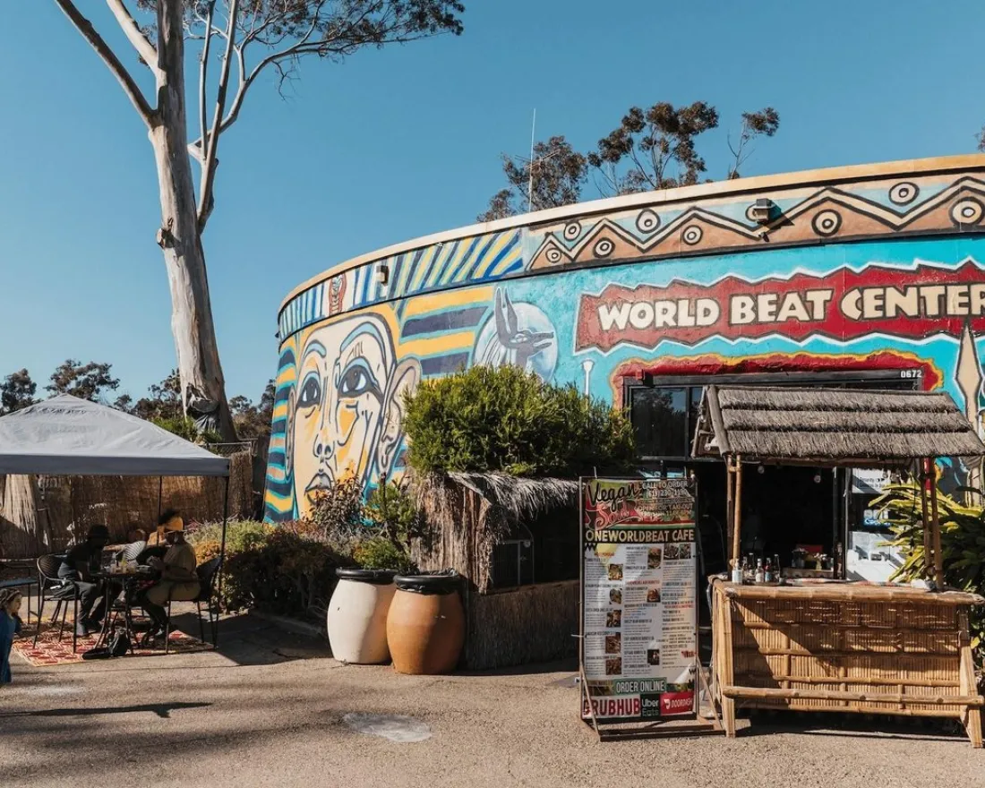 Outdoor view of the WorldBeat Center in Balboa Park, featuring a colorful mural, a small café stand, umbrellas, and people sitting and walking in the sun.