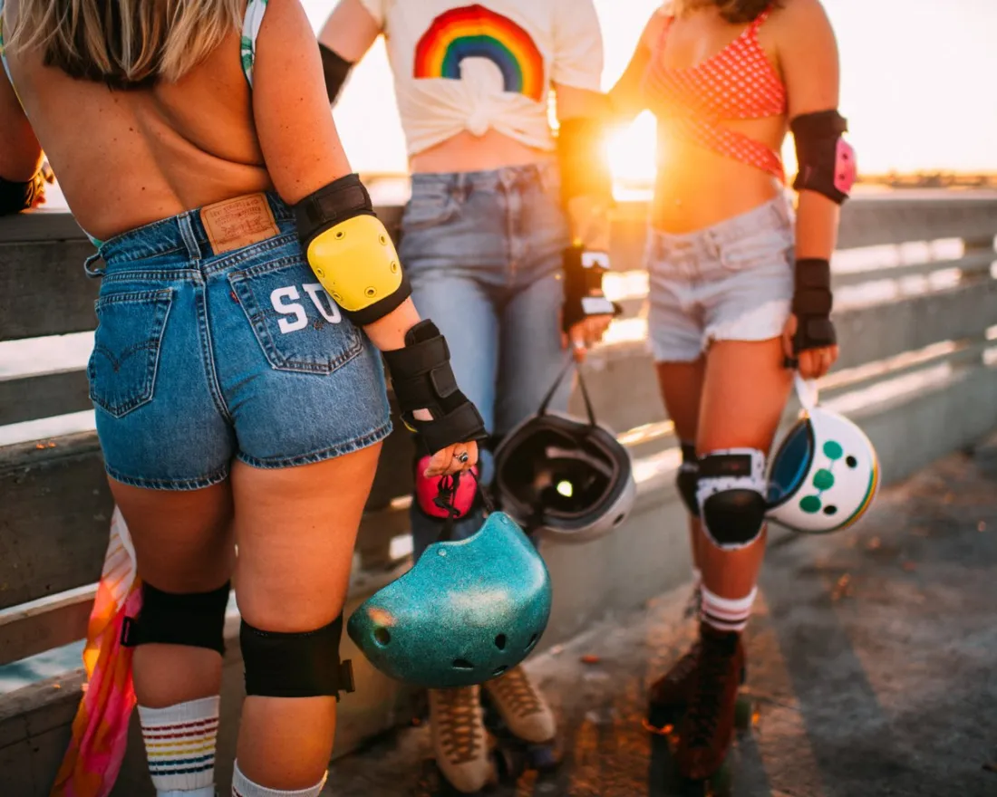 Three people in casual summer clothing and roller derby gear stand on a boardwalk at sunset, holding helmets and wearing knee and elbow pads.