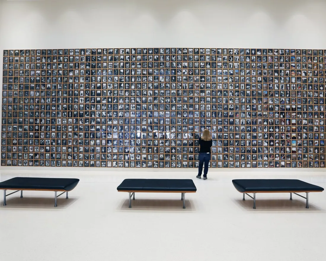 A person stands in front of a large wall display filled with many small framed photographs in a spacious, modern gallery with three black benches.