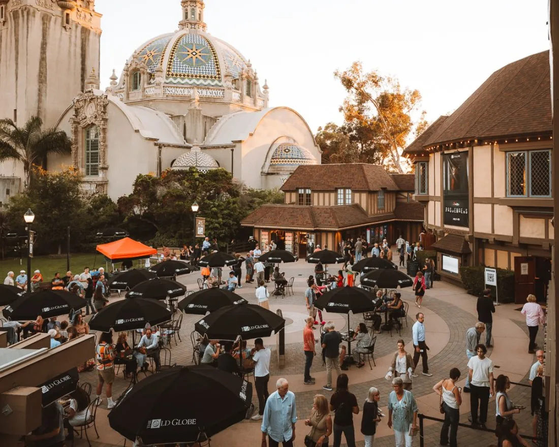 People gather in an outdoor courtyard with tables and umbrellas near historic buildings and a domed structure under a clear sky.