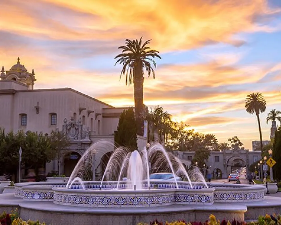 A fountain surrounded by flowers in front of historic Spanish-style buildings at sunset, with a tall tower and palm trees in the background.