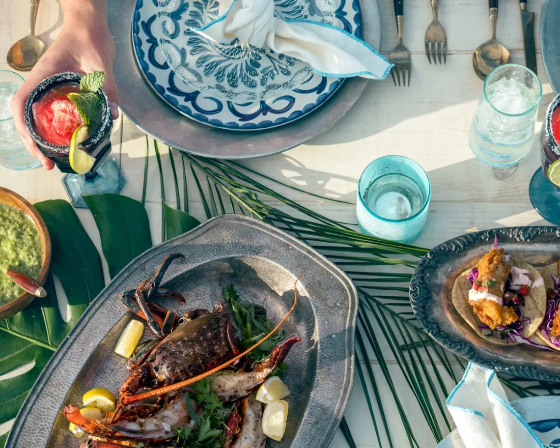 A table set with seafood, tacos, guacamole, blue and white plates, drinks with ice and lime, and cutlery, seen from above. A hand reaches for a red cocktail.
