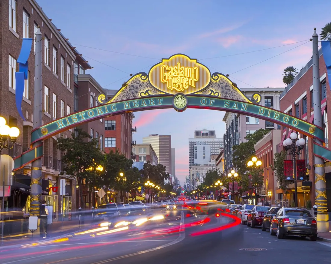 The entrance arch to San Diego’s Gaslamp Quarter is shown over a busy street with blurred car lights and historic buildings at dusk.