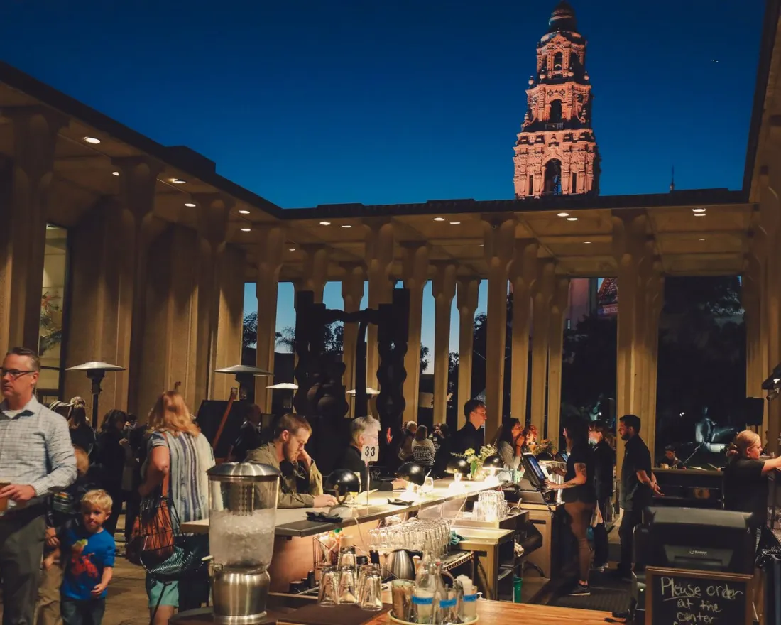 People gather at an outdoor bar and dining area in the evening; a tall illuminated tower is visible in the background against a dark blue sky.