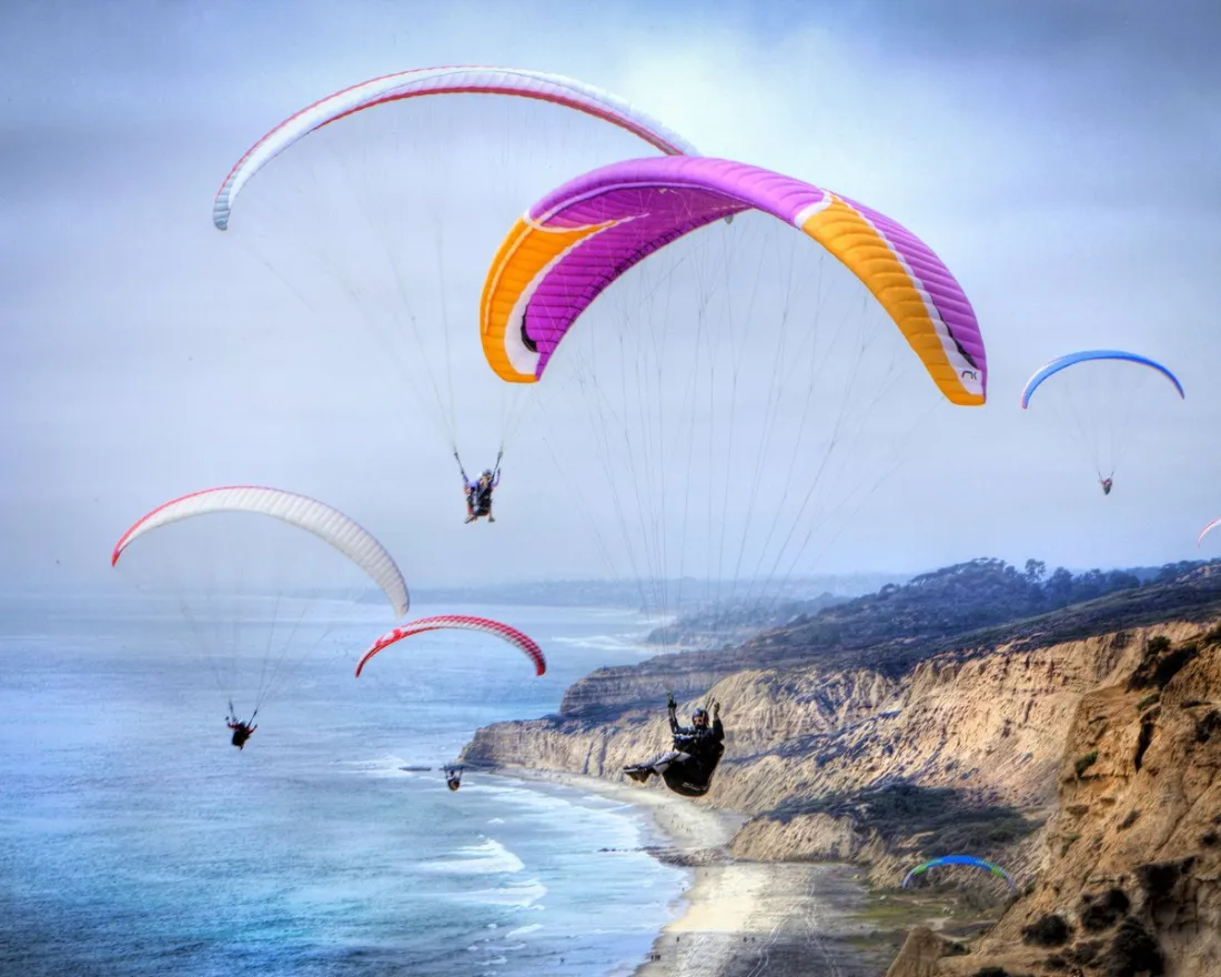 Several paragliders fly above coastal cliffs and the ocean under a cloudy sky, with a sandy shoreline and hilly landscape in the background.