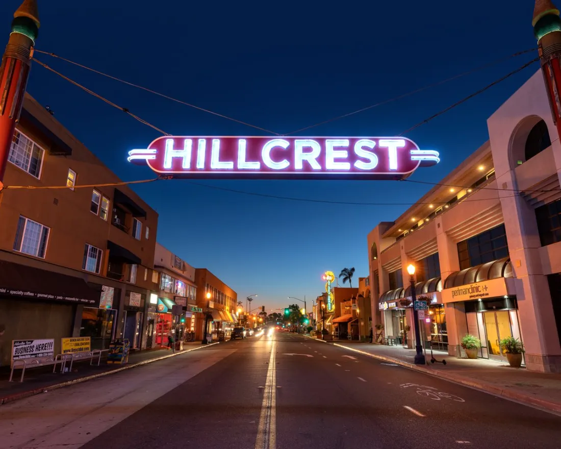 A neon "HILLCREST" sign hangs over a city street lined with shops and buildings at dusk, with streetlights and a clear evening sky.