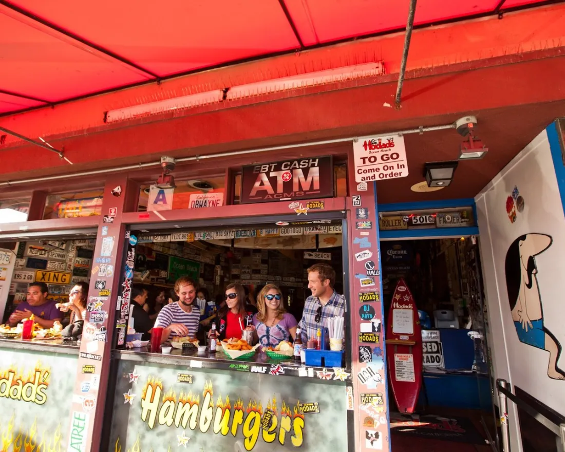 People sit at a counter eating food at an outdoor hamburger restaurant decorated with various signs and license plates under a red awning.