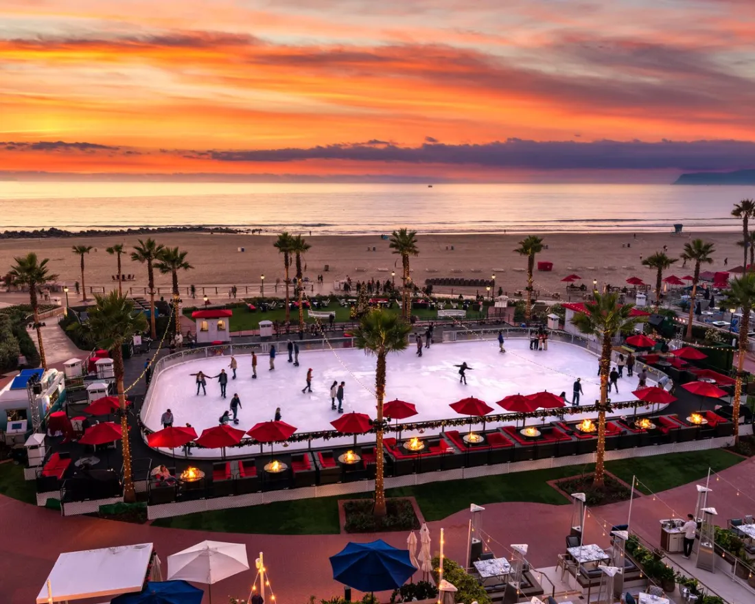 An outdoor ice skating rink near the beach is surrounded by palm trees and red umbrellas at sunset, with people skating and the ocean in the background.