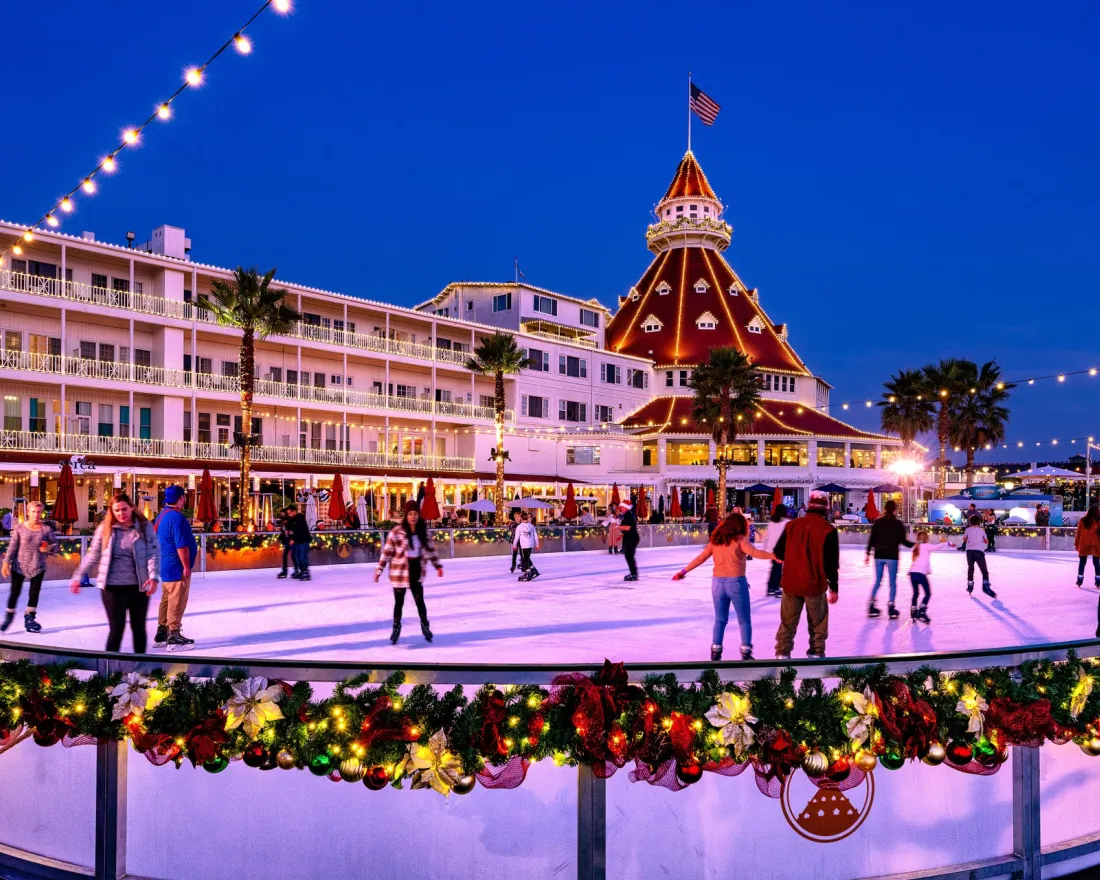 People ice skating on a decorated outdoor rink at dusk, with a large historic hotel and festive holiday lights in the background.
