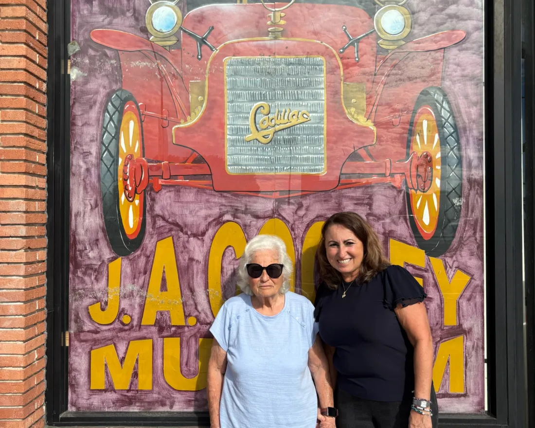 Two women stand smiling in front of a mural of a red antique car outside the J.A. Cooley Museum, which specializes in antique cars.