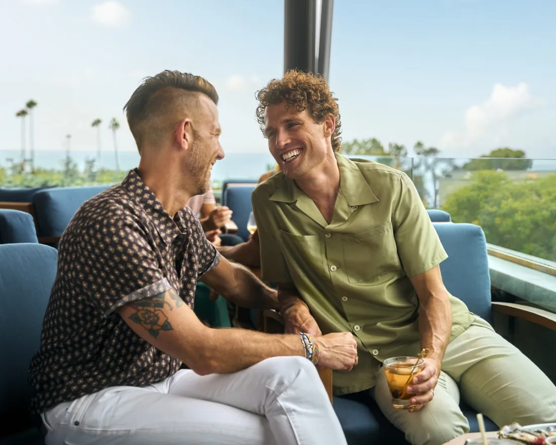 Two men sit on blue chairs at an outdoor lounge, smiling and holding drinks, with trees and a view of the ocean in the background.