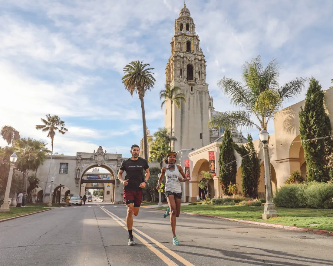 Two people run down a street lined with palm trees and historic buildings, with a tall tower in the background under a partly cloudy sky.