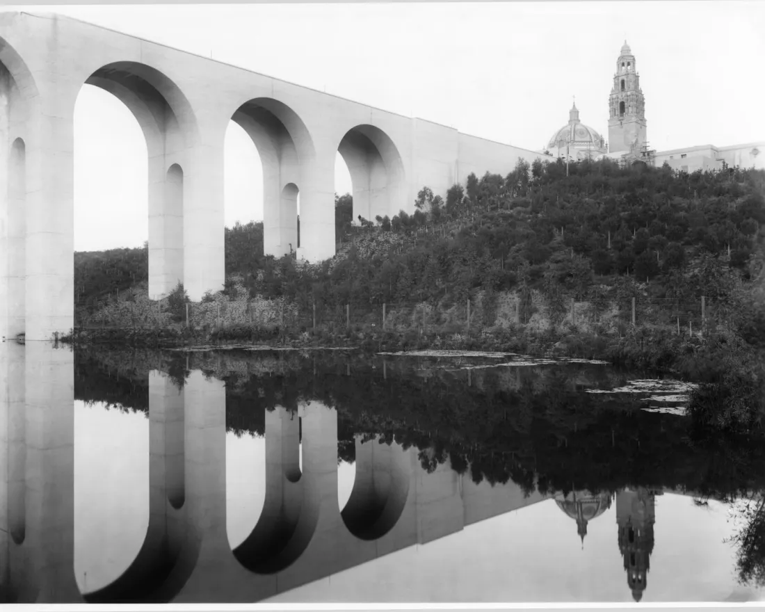 A large arched bridge spans over a river, reflecting in the water below, with a domed building and tower visible on a wooded hill in the background.