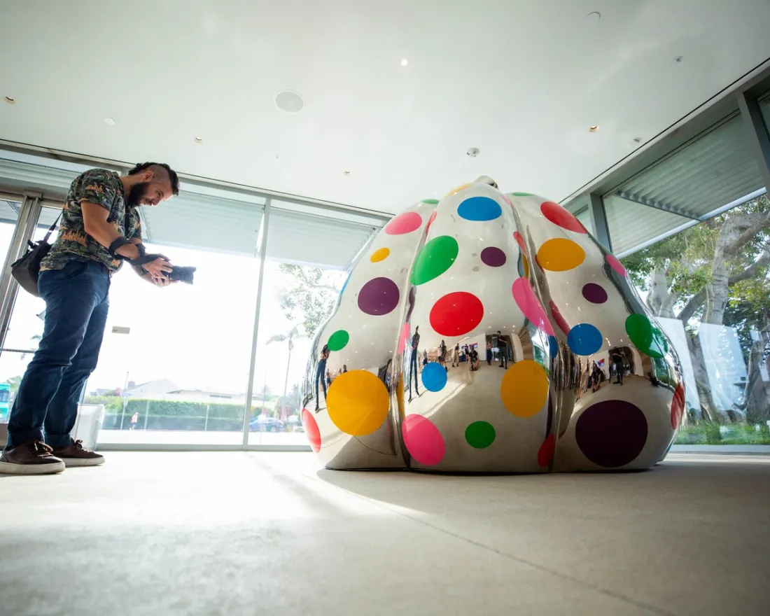 A man photographs a large, reflective sculpture covered in colorful polka dots inside a modern building with glass doors and windows.