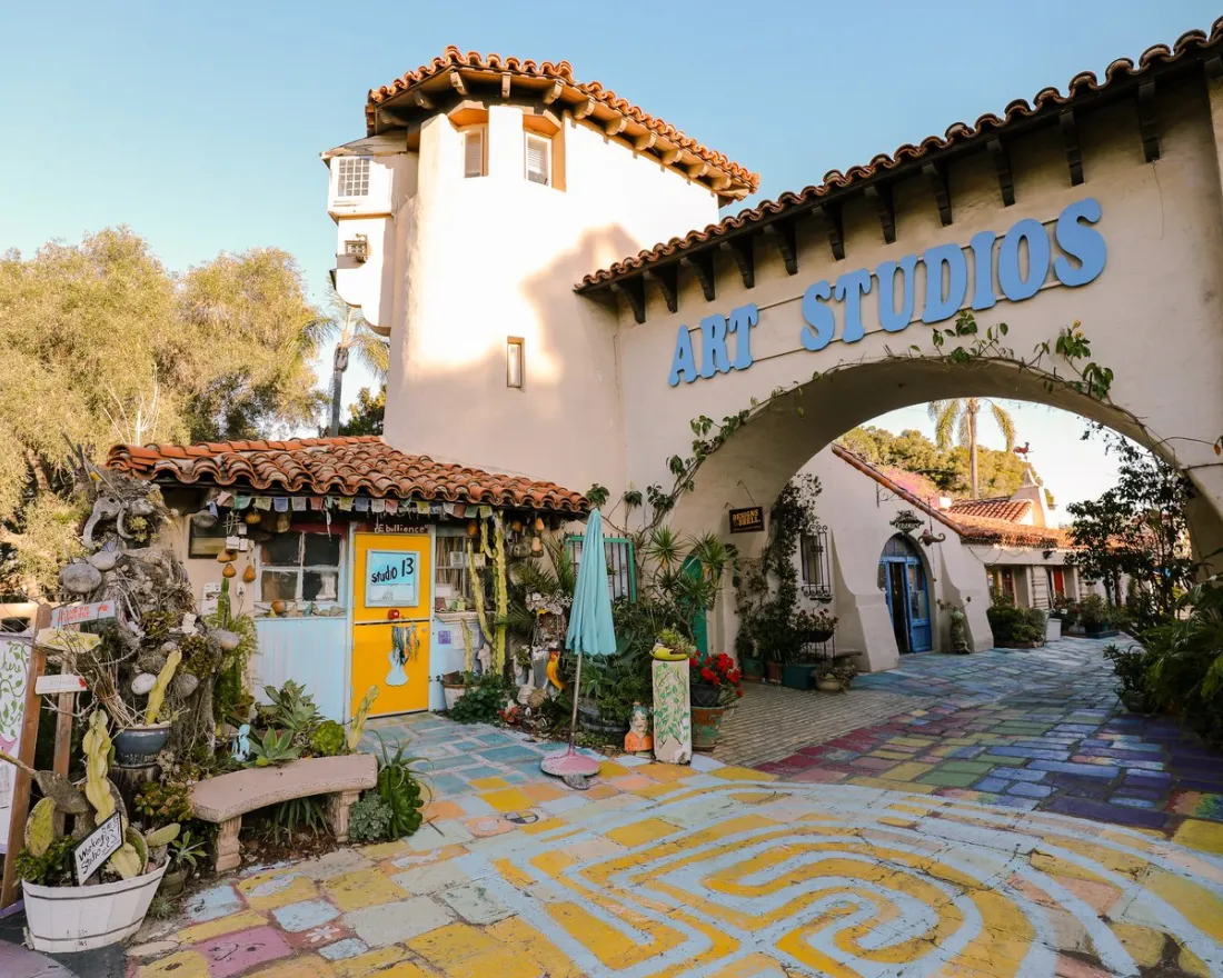 Colorful outdoor art studio with painted walkway, potted plants, tiled roofs, and a large “ART STUDIOS” sign above an archway on a sunny day.